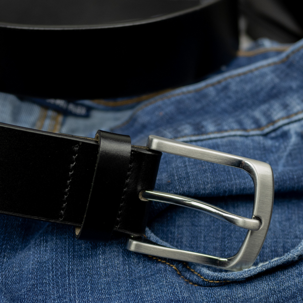 Black leather belt strap with curved low profile silver buckle on blue jean background. Nickel Free